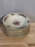 Stack of 12 ivory white vintage dinner plates with floral designs and gold scalloped edge, displayed on wooden surface.