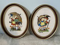 Photo of the set of two vintage Hummel needlepoint oval framed wall hangings side by side, showing detailed needlework of children holding umbrellas.