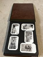 Brown cover photo album open to pages showing black and white photos held in photo corners on black pages. Photos depict buildings and outdoor scenes.