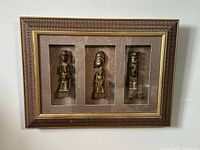 Front view of first vintage wood framed shadow box displaying three carved tribal/tiki style figures against a brown textured background. Frame features ornate wood carving with gold inner border.