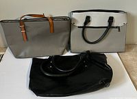 Photo of three tote bags side by side on a white surface showing beige bag with tan handles, white and grey bag with black handles, and large black bag.