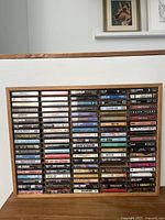 Wide view of wooden tape display holder filled with about 70 music cassette tapes arranged in rows, showing various artist names and album titles on spines.