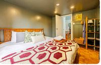 Bedroom showing king mattress with decorative pillows, cream and red crocheted coverlet, and wooden headboard.