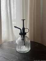 Clear ribbed glass plant mister with black pump and nozzle on dark wooden surface, neutral curtain background.