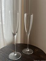 Pair of clear glass champagne flutes standing side-by-side on a dark wooden surface in front of light-colored curtains, showing the full glasses from base to rim.