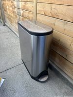 Closed view of stainless steel rectangular trash can with black trim and step pedal against wooden fence outdoors.