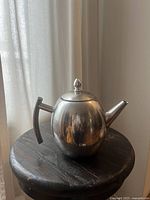 Silver-colored stainless steel teapot with vertical handle and spout placed on a round wooden surface against a curtain backdrop.