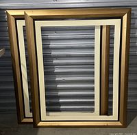 Two stacked large rectangular frames with gold edges and white inner borders leaning against a corrugated gray background.