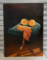 Front view of vintage oil painting showing half melon on blue cushioned stool with orange cloth draped over it, signed by artist