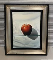 Front view of vintage framed painting showing a red apple against a white draped cloth background on a glass shelf.