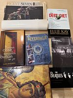 Books, DVD and newspaper displayed on table including Beatles book, Riverdale Student Handbook, Debt Diet by Ellie Kay, Benjamin Button, Chris Botti DVD, and vintage sports-focused newspaper.