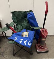 Two green folding camping chairs with armrests and metal legs, a red folding umbrella, a blue drawstring bag on one chair, and a red upright Daredevil vacuum cleaner standing beside them.