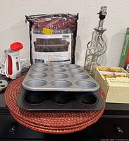 Photo showing velvet chair cover in packaging, two muffin pans stacked, red wicker placemats, Dolce wine bottle lamp, and box of gold flower floating candles.