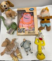 Disney plush toys and Monsters Inc. Sulley Blox figurine arranged on white cloth. Lady and the Tramp Lady plush, Elliott the dragon plush, Fantasia hippo plush, dwarf plush, Lumiere yellow candle plush, Mulan figurine, and Sulley Blox toy in box visible.