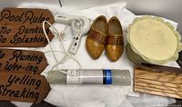 Photo of a vintage wooden pool rules sign, a white handheld hairdryer, vintage Dutch wooden clogs, a cream-colored cake plate, a wooden plank, and a rolled bath mat.