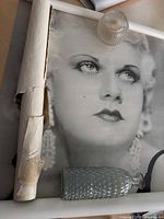 Photo of large black and white Mae West poster partially rolled and showing tears, with small clear textured glass vase and hobnail globe on top.