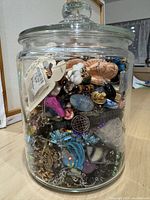 Glass jar filled with assorted jewelry components showing a variety of beads, charms, and chains in different colors and materials.