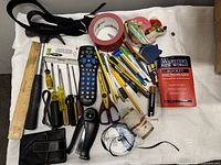 Full layout of assorted junk drawer items on white cloth showing tools, writing instruments, tape, wallet, stapler, remote, ruler, dictionary.