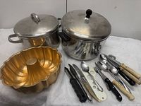 Overall view of two stainless steel pots with lids, copper bundt pan, assorted utensils with varied handles on white cloth