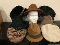 Front view of nine vintage men's hats including plaid fedoras, corduroy fedoras, flat caps, and faux fur hats around a mannequin head.
