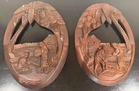 Full view of two oval Polynesian wood carving plaques showing village scenes with palm trees and huts, including man and woman figures.