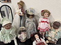 Wide view showing seven vintage porcelain dolls with detailed clothing, one doll inside glass dome, variety of hairstyles and skin tones