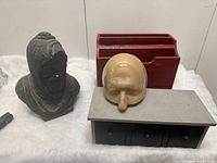 Three items displayed on white fabric: dark gray bust sculpture on the left, beige avant-garde head storage box front-center, and red vintage mail holder at the back.