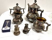 Partial silver plated tea set with teapot, sugar bowl with lid, creamer, and covered pot, pot belly stove shaped salt and pepper shakers, and boxed US Mint 50 State Quarter collectible spoon on white table.