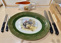 Turkey platter with green and gold decorative border and painted turkey centerpiece, accompanied by four knives, two large carving knives and two smaller knives with black handles.