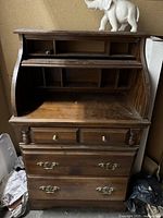 Front view of vintage wood secretary desk with roll-top open, two small drawers above and two larger drawers below, brass hardware visible, elephant statue on top.