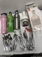 Overall view of water bottles, stainless steel utensils, and manual chopper with box and attachments on table.