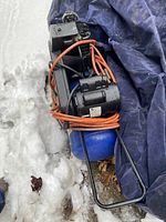Side view of blue air compressor with orange power cord on snowy ground with blue tarp behind.