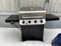 Char-Broil barbecue grill outdoors on snow with stainless steel lid and four control knobs, two black side shelves, and black base.