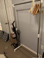 Metal clothes rack with multiple hangers, positioned against wall near vacuum cleaner and door.