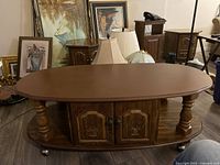Oval wooden coffee table with a polished wood top, carved floral design on the cabinet doors beneath, and turned legs on wheels.