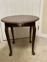 Full view of wooden oval-shaped end table with brass handle and four cabriole legs.