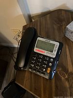 Black VTech digital desk phone on wooden surface showing keypad, buttons and digital display with date and caller ID information.