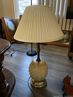 One photo showing a table lamp with a textured beige ceramic base, a dark wooden neck, a pleated cream-colored lampshade, and a clear acrylic bottom on a round metal stand.