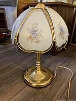 Table lamp with brass base and floral patterned glass shade, shown on wooden floor near wood furniture.