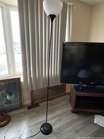 Tall floor lamp with black base and white opaque shade standing next to window and TV stand.