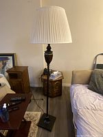 Photo showing a tall floor lamp with a white pleated shade and dark decorative metal base positioned next to a couch and side tables in a living room setting.