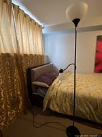 The photo shows a tall black floor lamp with a white bowl-shaped uplight and a smaller adjustable reading light beside a bed, situated in a bedroom with brown curtains and a bed with a yellow floral cover.