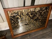 Photo of rectangular mirror with wooden frame and black-gold marbled pattern on reflective surface, placed against a wall.