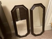 Two octagonal-shaped mirrors with dark brown wooden frames leaning against a beige wall and beige carpet floor.