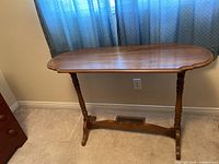 Photo showing a wooden side table with a medium brown finish, two turned spindle-style legs, a stretcher connecting the legs at the base, placed against a wall under a window with blue curtains.