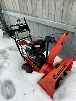 Front-angle view showing orange steel auger housing and black plastic discharge chute in snow