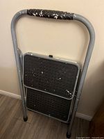 Front view of gray metal 2-step stool showing wear and paint chipping on frame and textured black steps.