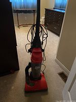 Photo of red Dirt Devil Featherlite upright vacuum cleaner with cord wrapped on holder and flexible hose attached, placed on carpet in a room with furniture.