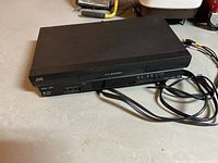 Black JVC HR-J692U VCR player with power cord and RCA audio/video cables visible on concrete surface.