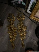 Two ornate gold-tone wall candle holders placed on a wooden floor, showing intricate floral and ribbon designs.